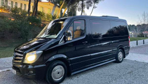A black Mercedes Sprinter minibus parked at dusk, reflecting the warm hues of the setting sun.
